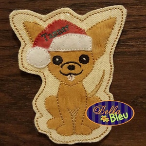 Puede incluir: Un chihuahua marrón y blanco con un gorro de Papá Noel rojo y blanco con el nombre "Tanner" bordado. El chihuahua está sentado con la lengua fuera. La imagen está sobre un fondo de tela beige con un borde bordado dorado.