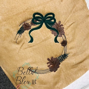 Puede incluir: Manta beige con un diseño de corona bordada. La corona presenta piñas, vegetación y un lazo verde oscuro. La manta tiene un borde blanco y esponjoso y el texto "Bella Bleu LLC".