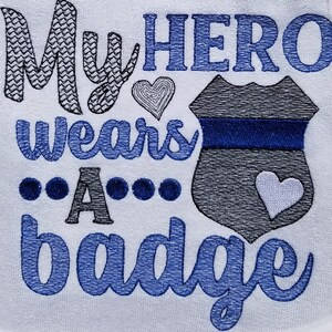 May include: Embroidered white towel with blue and gray stitching. The text reads "My HERO wears A badge" with a heart and a police badge design.