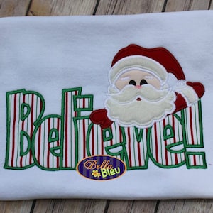 May include: White t-shirt with the word "BELIEVE" in green and red striped letters, and a Santa Claus applique. The Santa has a red hat and white beard. A small oval logo with the text "Bella Bleu" is at the bottom.