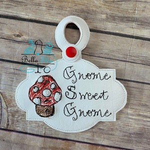 Może przedstawiać: Biały filcowy wieszak na drzwi w kształcie gnoma z czerwonym guzikiem i wzorem grzyba. Tekst "Gnome Sweet Gnome" jest wyszyty na wieszaku.