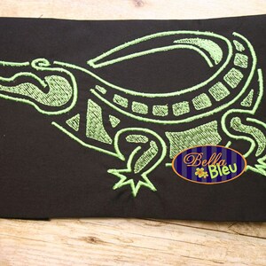 Könnte beinhalten: Grüne gestickte Alligator-Darstellung auf schwarzem Stoffhintergrund. Das Design ist stilisiert und weist ein geometrisches Muster auf. Der Text "Bella Bleu" ist in einem weißen Oval mit Blumenmuster sichtbar.