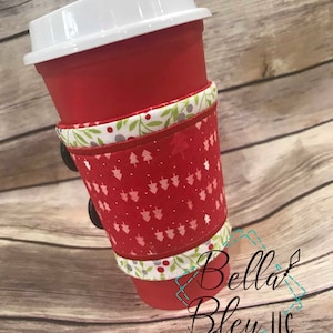 May include: Red and white Christmas-themed fabric coffee cup cozy with a green and white trim. The cozy is on a red to-go cup with a white lid. The text "Bella Bleu LLC" is visible in the bottom right corner of the image.