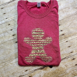 May include: A folded maroon t-shirt with a gingerbread man design. The gingerbread man is made of a leopard print pattern with horizontal stripes. The shirt is on a wooden surface.
