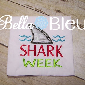 May include: White t-shirt with a grey shark fin, blue waves, and the text "SHARK WEEK" in red and green.
