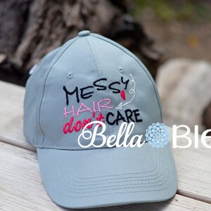May include: A light gray baseball cap with the embroidered text "Messy Hair don't care" in black, pink, and red thread. The text is surrounded by a white heart and a white arrow. The cap has a black bill and a white logo that says "Bella Bleu".