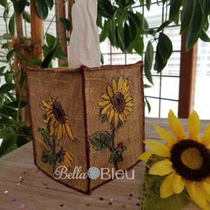 Puede incluir: Caja de pañuelos rectangular decorada con girasoles bordados sobre tela tipo arpillera. La caja tiene un diseño de girasol en cada lado. El texto "Bella Bleu" es visible en la parte delantera.