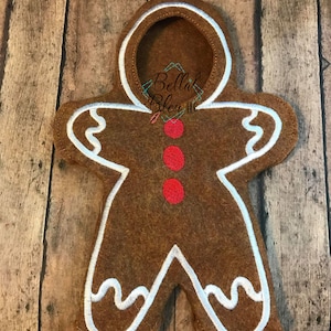 May include: A brown felt gingerbread man cutout with white icing details and red buttons. The gingerbread man has a hole for a head.