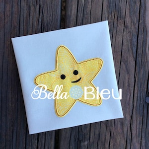 May include: A yellow embroidered star with a smiling face on a white fabric background. The text "Bella Bleu" is visible below the star.