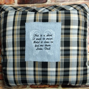 Pode incluir: Uma almofada de xadrez preto e branco com um remendo de tecido azul claro costurado na frente. O remendo de tecido tem uma mensagem bordada que diz "This is a shirt I used to wear. Hold it close to feel me there. Love, Dad." A mensagem é cercada por asas bordadas.