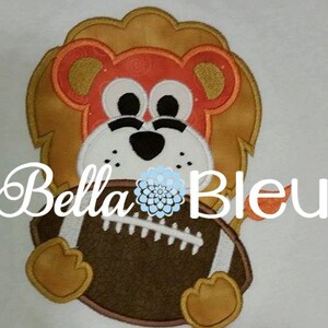 May include: Embroidered applique of a brown lion holding a brown and white football. The lion has a light orange face with black eyes and a black nose.