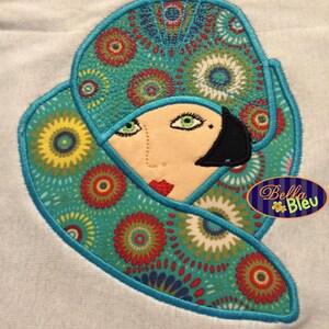 May include: Embroidered design of a woman wearing a teal and red patterned hat and scarf. The design is outlined in teal thread and features a black eye and a red mouth.