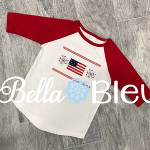 May include: White and red baseball tee shirt with a red, white, and blue American flag and fireworks design. The shirt has a red raglan sleeve.