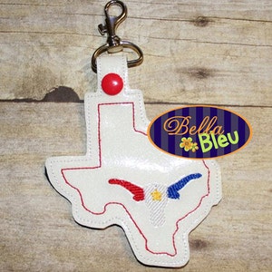 May include: A white leather keychain shaped like the state of Texas with a red, white, and blue longhorn steer design. The keychain has a silver metal clasp.