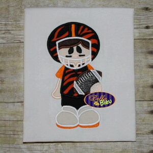 May include: Embroidered design of a cartoon football player wearing an orange and black striped jersey and helmet, holding a football.