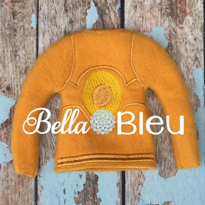 May include: A yellow felt elf sweater with a yellow embroidered sun design on the back. The sweater has a brown trim around the neck, cuffs, and hem. The text "Bella Bleu" is embroidered on the front of the sweater.
