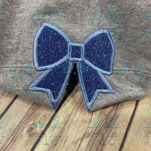 Könnte beinhalten: Eine blaue Glitzer Schleife mit einem hellblauen Umriss. Die Schleife ist auf einen grauen Stoff genäht.