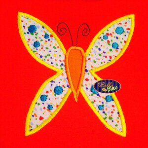 May include: A colourful butterfly applique with yellow, white, orange and blue accents. The butterfly is embroidered on a red fabric background. The text "Bella Bleu" is visible in the bottom right corner of the image.