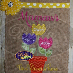 May include: Burlap garden flag with embroidered floral design. Features the text "Mamaw's" above three flower-shaped name tags: "Owen", "Sophia", and "Abby". A yellow flower and the words "love blooms here" are also included.