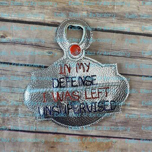 May include: A silver-colored, metallic,  round,  embellished patch with the text "In My Defense I Was Left Unsupervised". The text is in red, white, and blue thread.