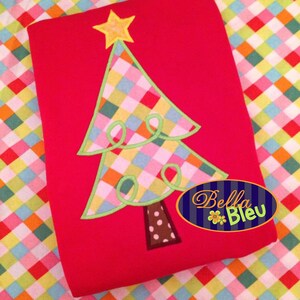 Pode incluir: Um tecido vermelho com uma aplicação de árvore de Natal xadrez colorida. A árvore tem uma estrela amarela no topo e um tronco marrom com bolinhas brancas. O tecido tem o texto "Bella Bleu" em um círculo azul com uma flor branca.