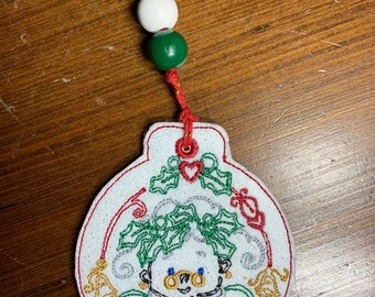May include: White felt Christmas ornament with a red, green, and gold embroidered design of a woman with holly leaves and a bird. The ornament has a red and gold string with three beads.