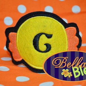 May include: A yellow felt candy applique with a black felt letter G in the centre. The candy is surrounded by orange felt with a scalloped edge. The applique is on a white and orange polka dot background. Bella Bleu is written in a white font on a blue background.