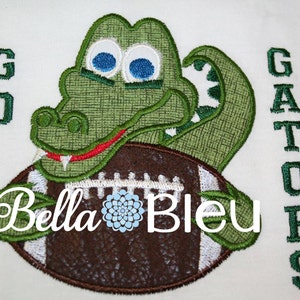 May include: Green alligator holding a brown and white football with the text "GO GATORS" embroidered on a white background.