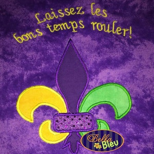 May include: Purple fabric with a gold, green, and purple fleur-de-lis embroidered design. The text "Laissez les bons temps rouler!" is embroidered in yellow above the fleur-de-lis.
