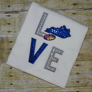Puede incluir: Camiseta blanca con la palabra "LOVE" en un diseño azul y plateado. La "O" se reemplaza con un contorno de lentejuelas azules del estado de Kentucky. La camiseta también presenta el logotipo "Bella Bleu".