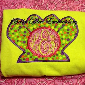 May include: A bright yellow t-shirt with a colorful embroidered design of a seashell with a circle in the center. The circle has a pink swirl design and the text "Bella Bleu Embroidery" is above the seashell.