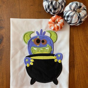 May include: A white kitchen towel featuring an embroidered design of a purple monster holding a black cauldron. The monster has green horns, yellow eyes, and a green mouth. The cauldron has a yellow rim and yellow dots on the sides.