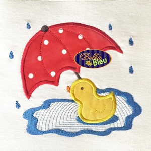 Puede incluir: Un pato de goma amarillo con una sombrilla roja con lunares blancos sobre él, sentado en un charco azul. La sombrilla tiene el texto "Bella Bleu".