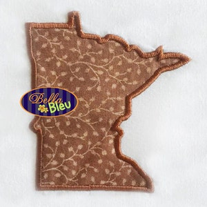 May include: A brown fabric applique of the state of Minnesota with a brown floral pattern and a brown satin stitch outline. The applique is on a white background. The text "Bella Bleu" is visible in the lower left corner of the image.
