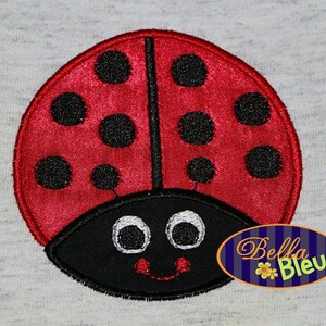 May include: Red ladybug with black spots and a black head with white eyes and a smile. The ladybug is embroidered on a gray fabric.