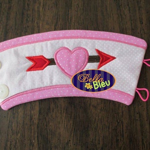 Op de afbeelding: Roze en witte stoffen koffiebekerhoes met een roze hart- en pijlontwerp. De hoes heeft twee lussen om aan een beker te bevestigen. De tekst "Bella Bleu" is geborduurd op de hoes.