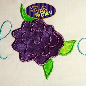 Könnte beinhalten: Stickerei mit den Buchstaben "H" in einer kursiven Schriftart, einer lila Rose mit grünen Blättern und dem Text "Bella Bleu".