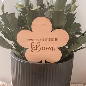 以下が含まれることがあります： 「私を咲かせてくれてありがとう」と書かれた木製のフラワー型の植物マーカー。マーカーは黒い鉢に植えられた緑色のサボテンの植物に入っています。
