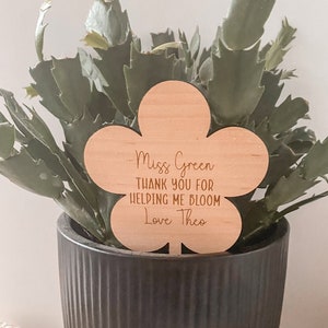 May include: A wooden flower-shaped plant marker with the text "Miss Green Thank you for helping me bloom Love Theo" engraved on it. The marker is placed in a black pot with a green plant.