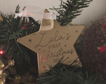 Könnte beinhalten: Goldener sternförmiger Weihnachtsbaumschmuck mit dem Text "Lola's First Christmas 2019" darauf.