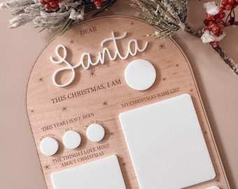 May include: A wooden Christmas countdown board with a white circle, a white rectangle, and three smaller white circles. The board has the text "Dear Santa", "This Christmas, I am", "This year I have been", "Naughty", "Nice", "A bit of both", "The things I love most about Christmas", "My Christmas wish list", and "Love from" written on it.