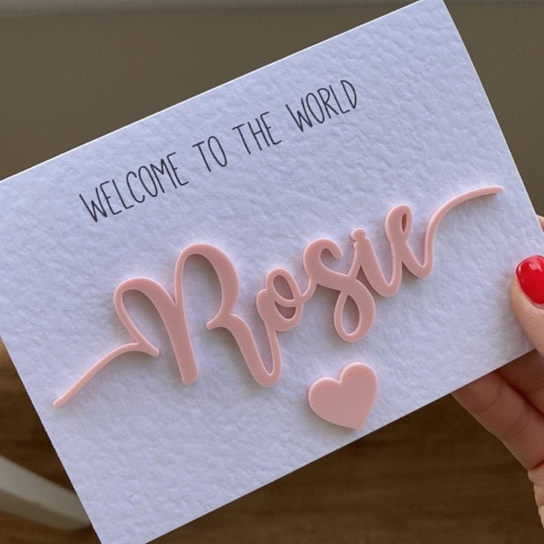 May include: A pink card with the text "WELCOME TO THE WORLD" and the name "Rosie" written in a cursive font. A pink heart is below the name.