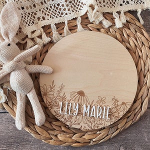 May include: A round wooden plaque with a floral design and the name "Lily Marie" engraved on it. The plaque is sitting on a woven wicker mat.