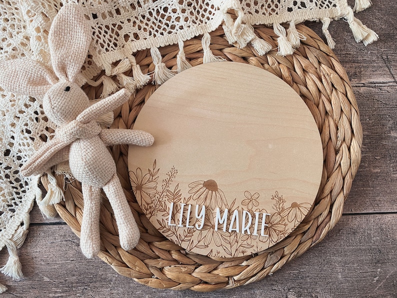 May include: A round wooden plaque with a floral design and the name "Lily Marie" engraved on it. The plaque is sitting on a woven wicker mat.