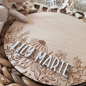 May include: A round wooden sign with a floral design engraved on the bottom half. The top half is blank and has the name "Lily Marie" spelled out in white letters.