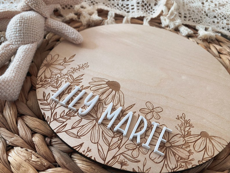 May include: A round wooden sign with a floral design engraved on the bottom half. The top half is blank and has the name "Lily Marie" spelled out in white letters.