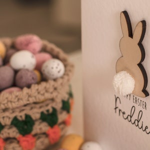 K&ouml;nnte beinhalten: Eine wei&szlig;e Karte mit einem braunen Hasen-Ausschnitt mit einem wei&szlig;en Bommel-Schwanz. Die Karte sagt "Frohe Ostern Freddie". Im Hintergrund befindet sich ein geh&auml;kelter Korb gef&uuml;llt mit Schokoladeneiern.