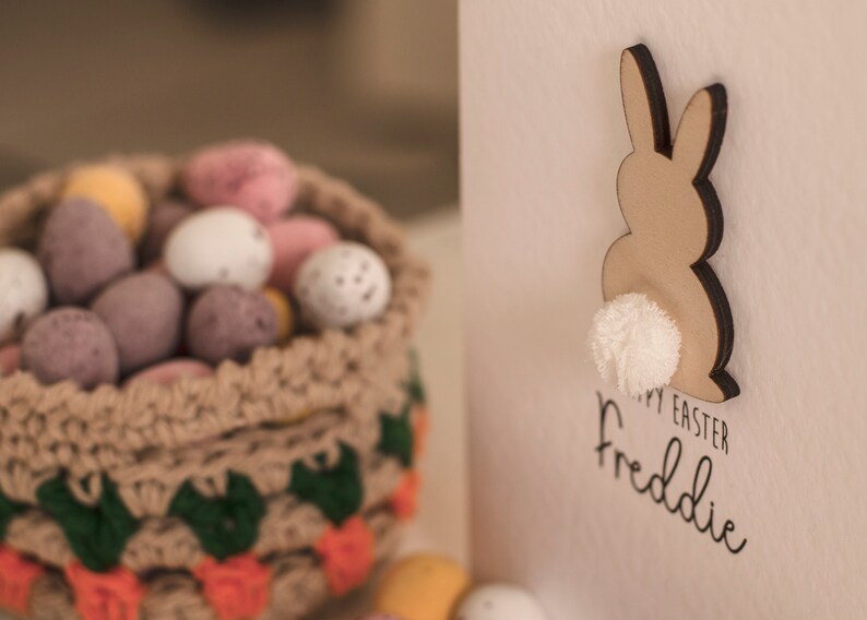 K&ouml;nnte beinhalten: Eine wei&szlig;e Karte mit einem braunen Hasen-Ausschnitt mit einem wei&szlig;en Bommel-Schwanz. Die Karte sagt "Frohe Ostern Freddie". Im Hintergrund befindet sich ein geh&auml;kelter Korb gef&uuml;llt mit Schokoladeneiern.