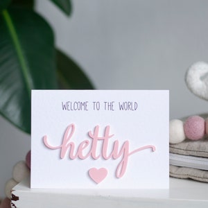 May include: A white card with the text "Welcome to the world" and the name "Hetty" written in pink 3D letters. A pink heart is below the name.