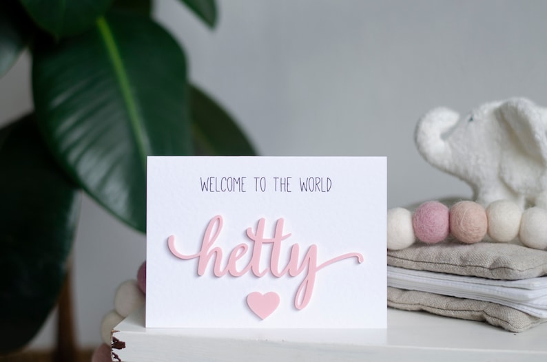 May include: A white card with the text "Welcome to the world" and the name "Hetty" written in pink 3D letters. A pink heart is below the name.